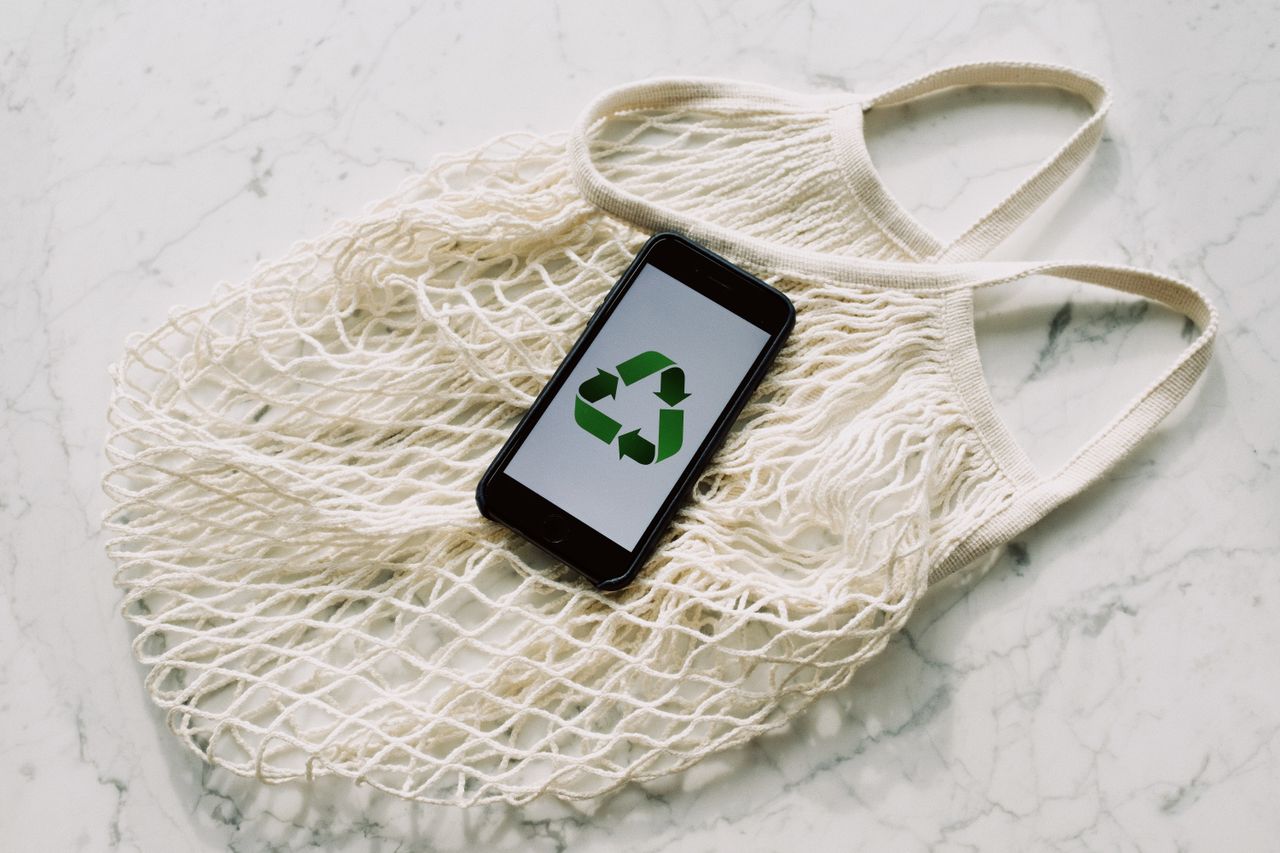 about-01 Overhead of smartphone with simple recycling sign on screen placed on white eco friendly mesh bag on marble table in room